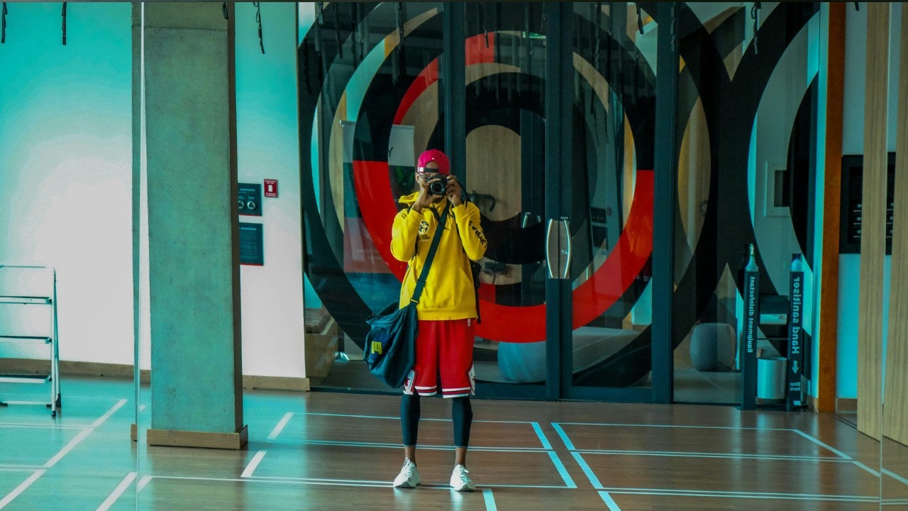 A photographer dressed in brightly colored clothes holds his camera to his eye in a lobby. 
