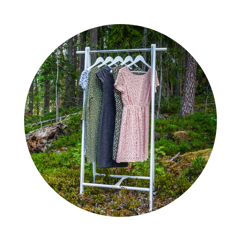 Ruhák fehér állványon, erdei környezetben felakasztva, természetközeli és fenntartható divat hangulatot idézve.