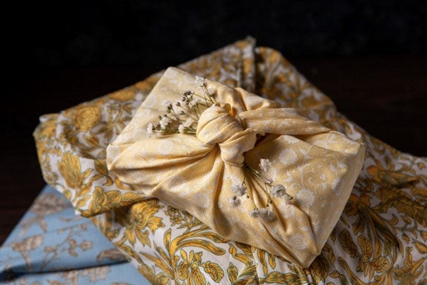 Textilbe csomagolt ajándék (furoshiki stílus), virágmintás anyaggal átkötve, természetes dekorációval díszítve.