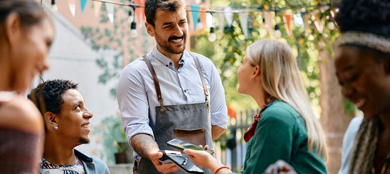 Waiter holding a payment terminal as a woman pays using her smartphone at an outdoor restaurant table with friends.