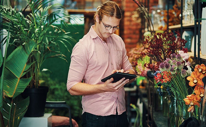 A man stands in a flower shop, focused on a tablet while surrounded by colorful floral arrangements.