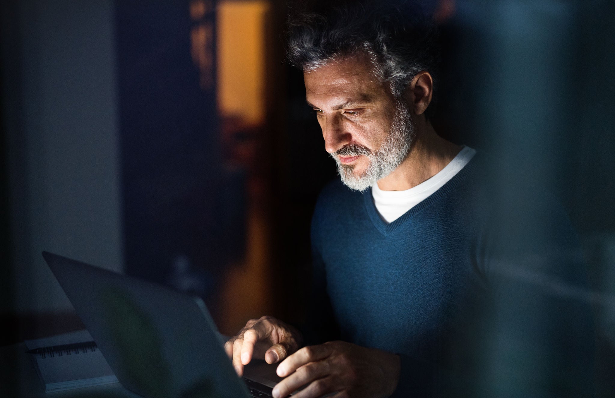 Handsome man working from home office at evening, using laptop. Screen glow illuminates man's face. Remote work.