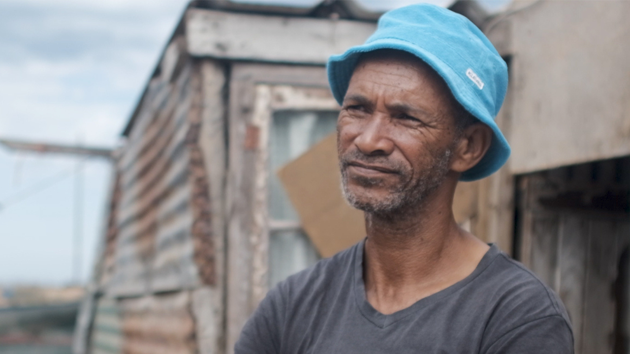 კაცი ლურჯი ვედრო ქუდით და ნაცრისფერი მაისურით დგას დგომებზე თუნხით დაფარული შენობის წინ.