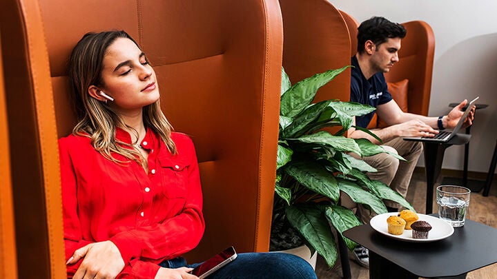 Fiatal nő kényelmes fülkében pihen okostelefonnal a kezében, mögötte fiatal férfi dolgozik laptopon kávézó jellegű belső térben