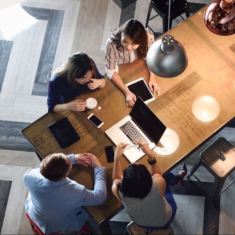 Several people sit at a table around a laptop talking in an office setting. 