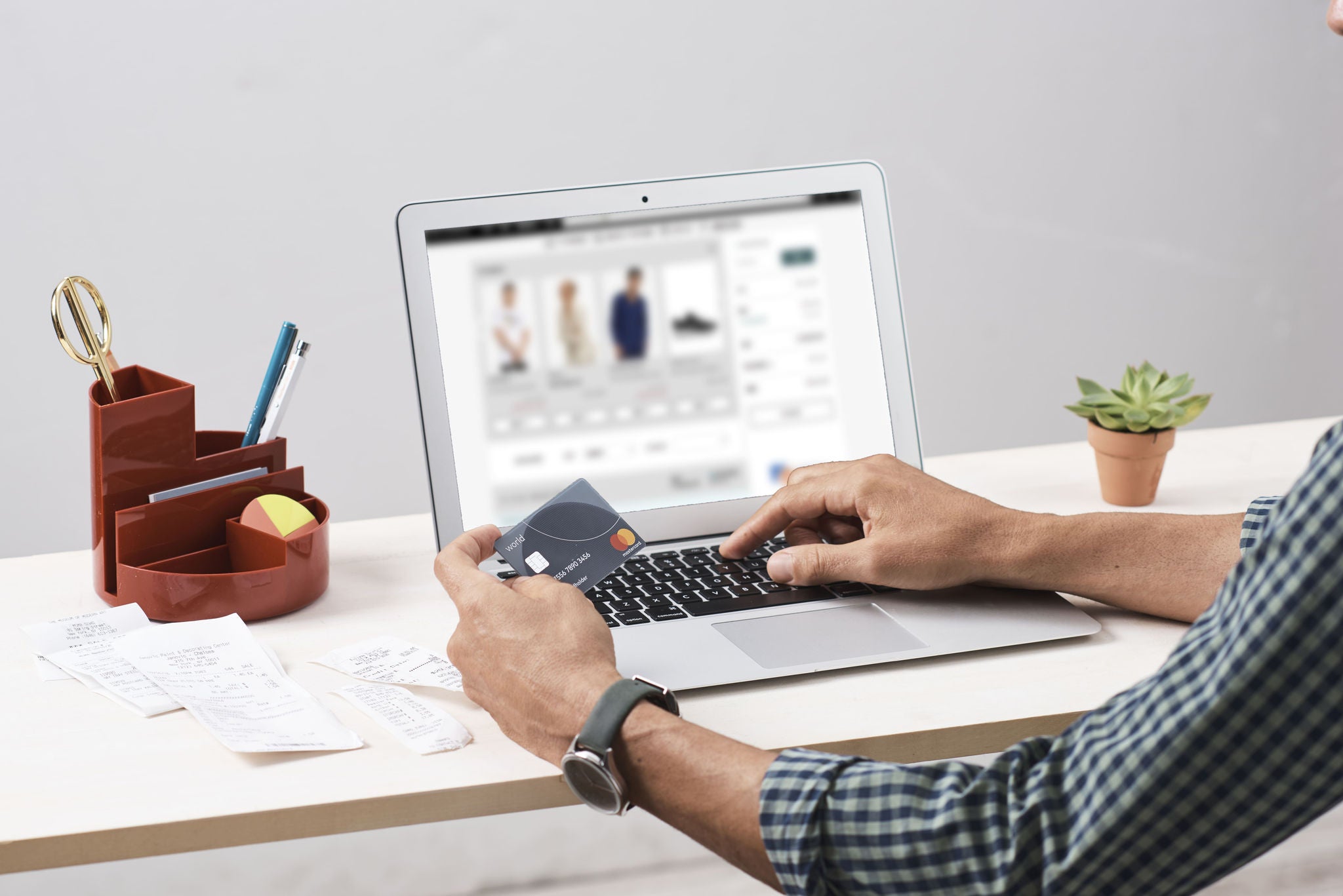 A man in a checked shirt sits at a desk holding a payment card while typing on a laptop showing an online clothing store, with receipts and a small plant nearby.