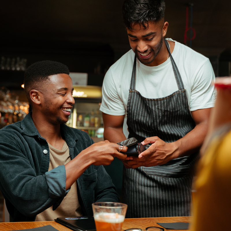 A man holds his phone to a waiter in an apron to pay his bill at a restaurant. 
