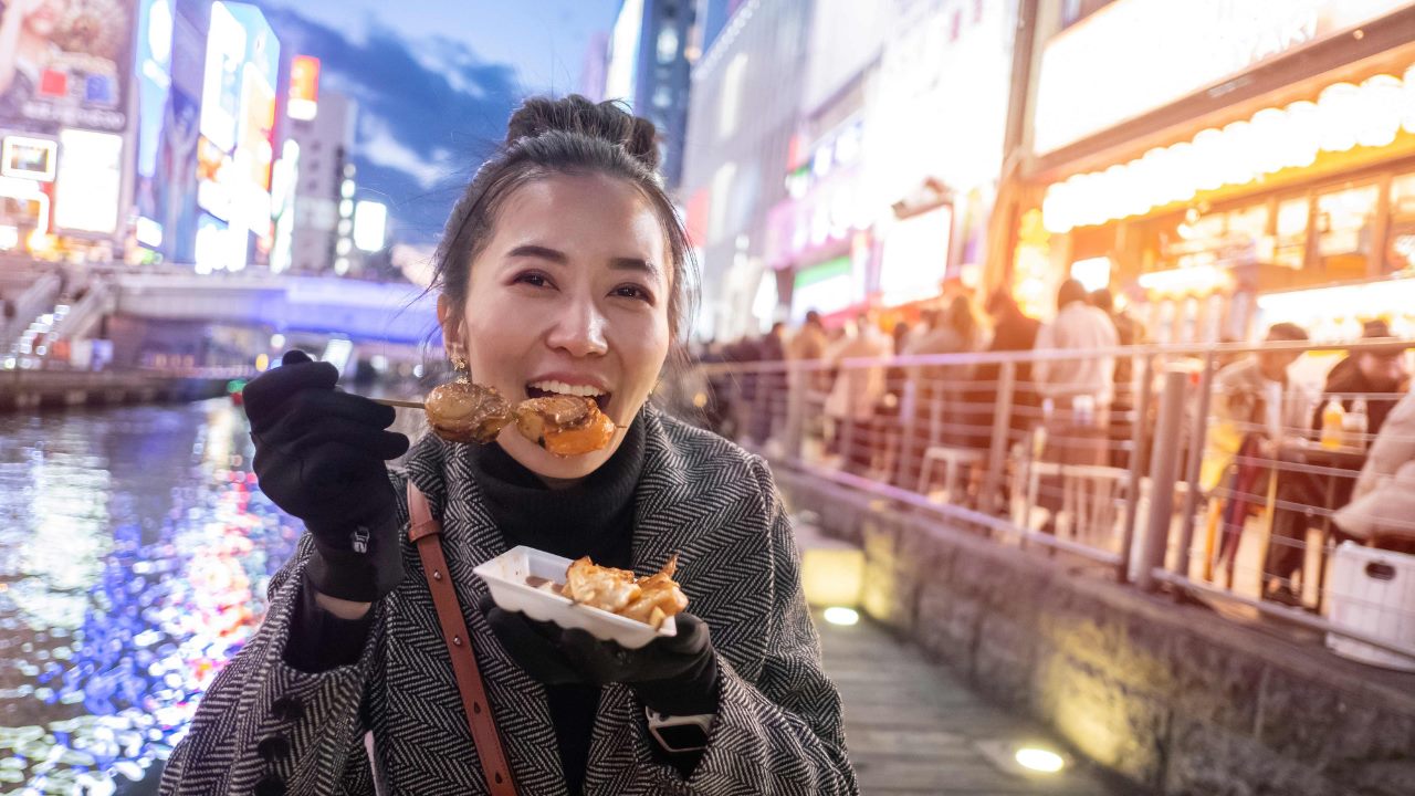 A woman eats fried octopus pancakes in Osaka next to a canal at night. 