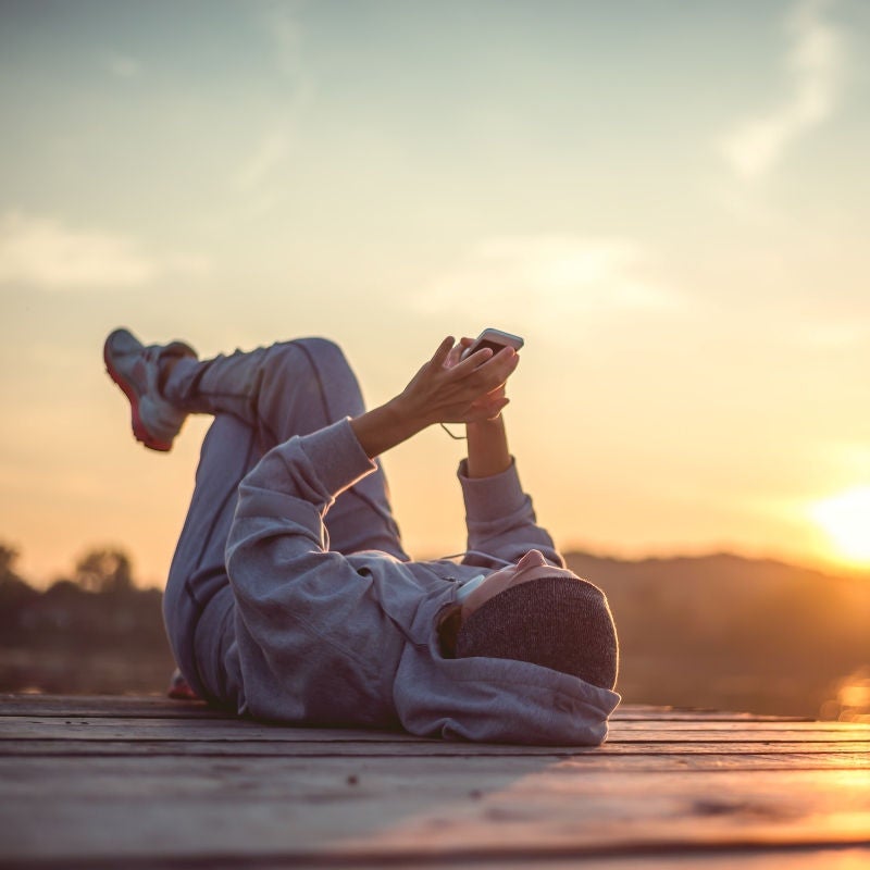 A woman lays on a dock in a running gear holding her phone as the sun rises. 
