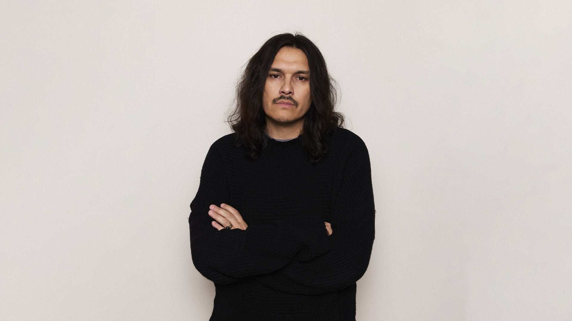 Portrait of fashion designer Guillermo Andrade standing with arms crossed, wearing a black textured sweater, against a plain off-white background.