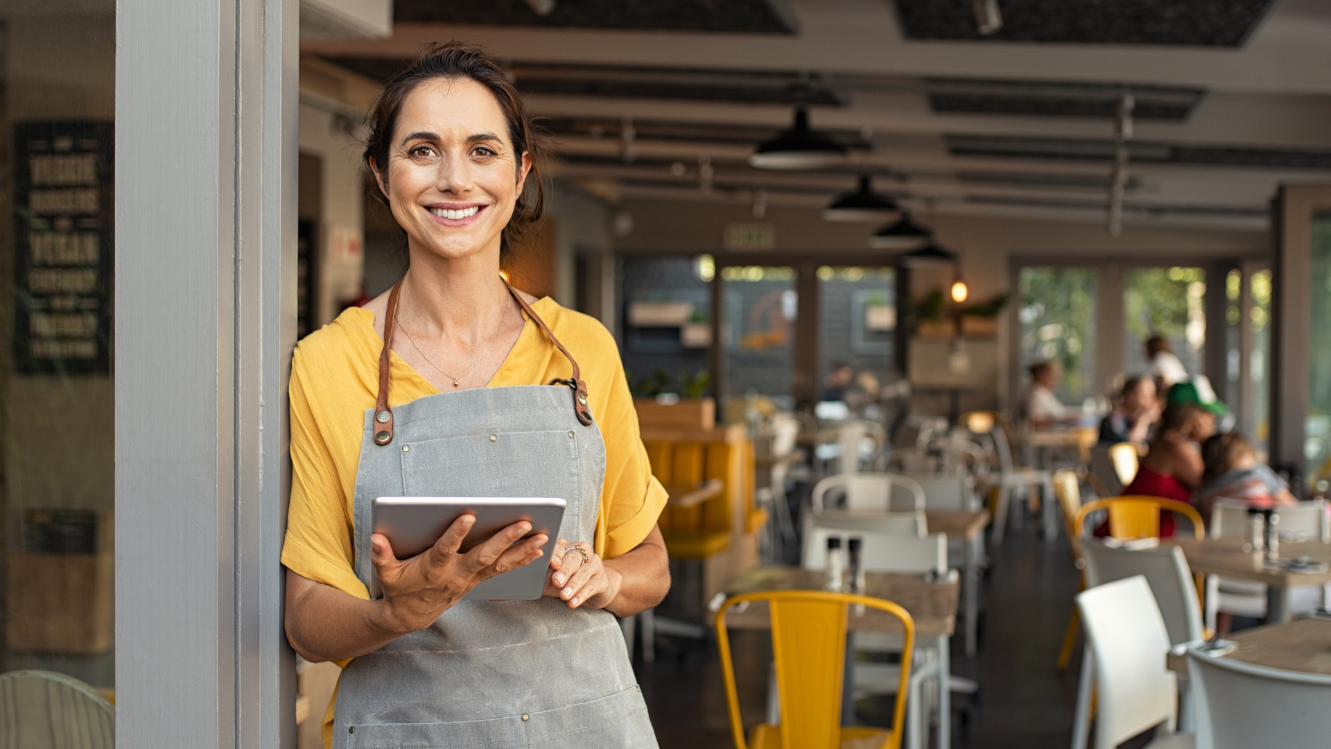 A woman smiles while holding a tablet in a café with chairs and people in the background.