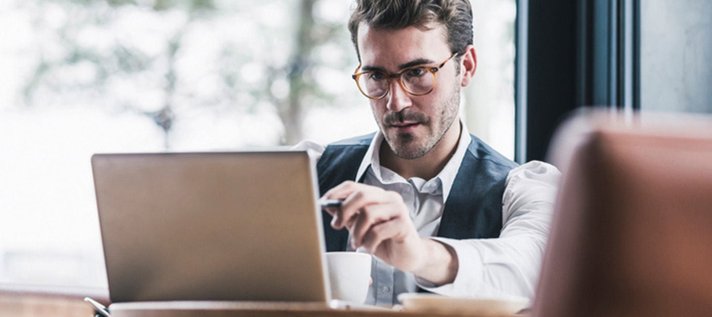 En person iført briller og vest, jobber konsentrert på sin bærbare datamaskin i kafé eller restaurantmiljø.