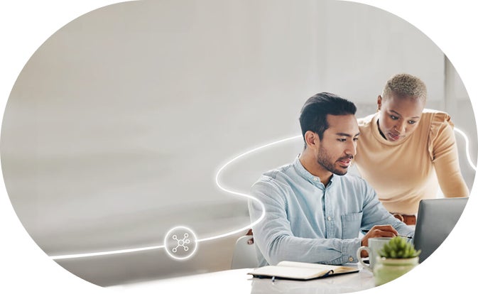 Two people work together at a desk, looking at a laptop screen.