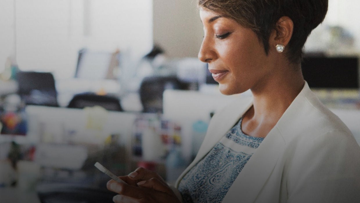 A woman in an office setting, focused on her phone while seated at her desk.