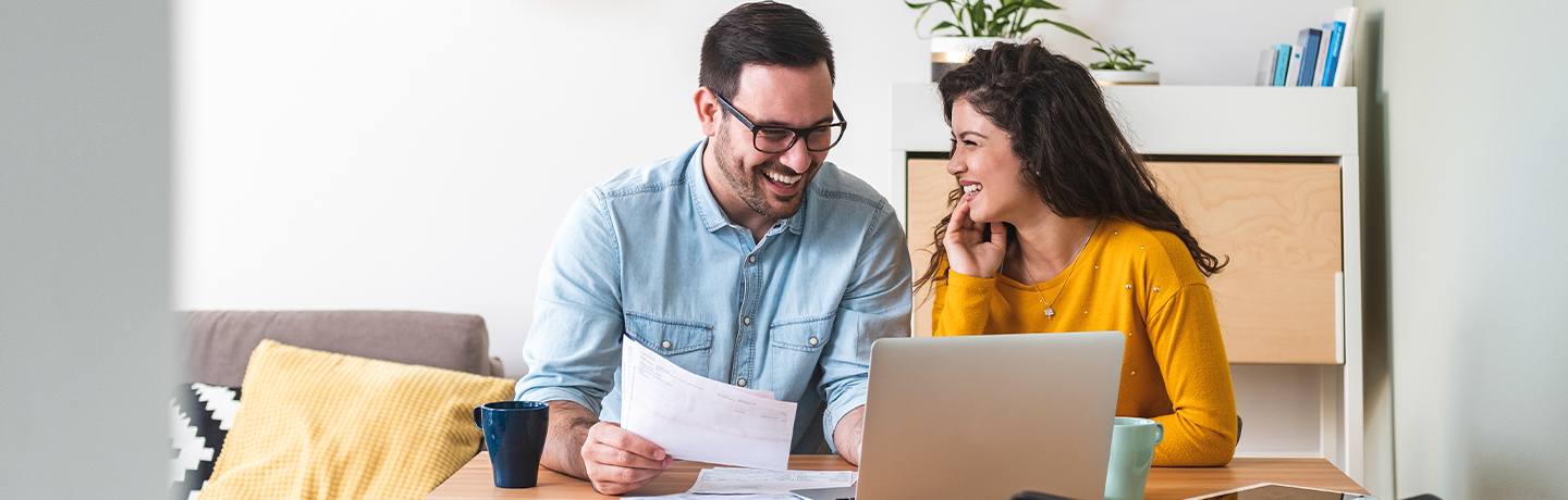 A man and a woman sitting on a sofa smiling at each other with a piece of paper and a laptop