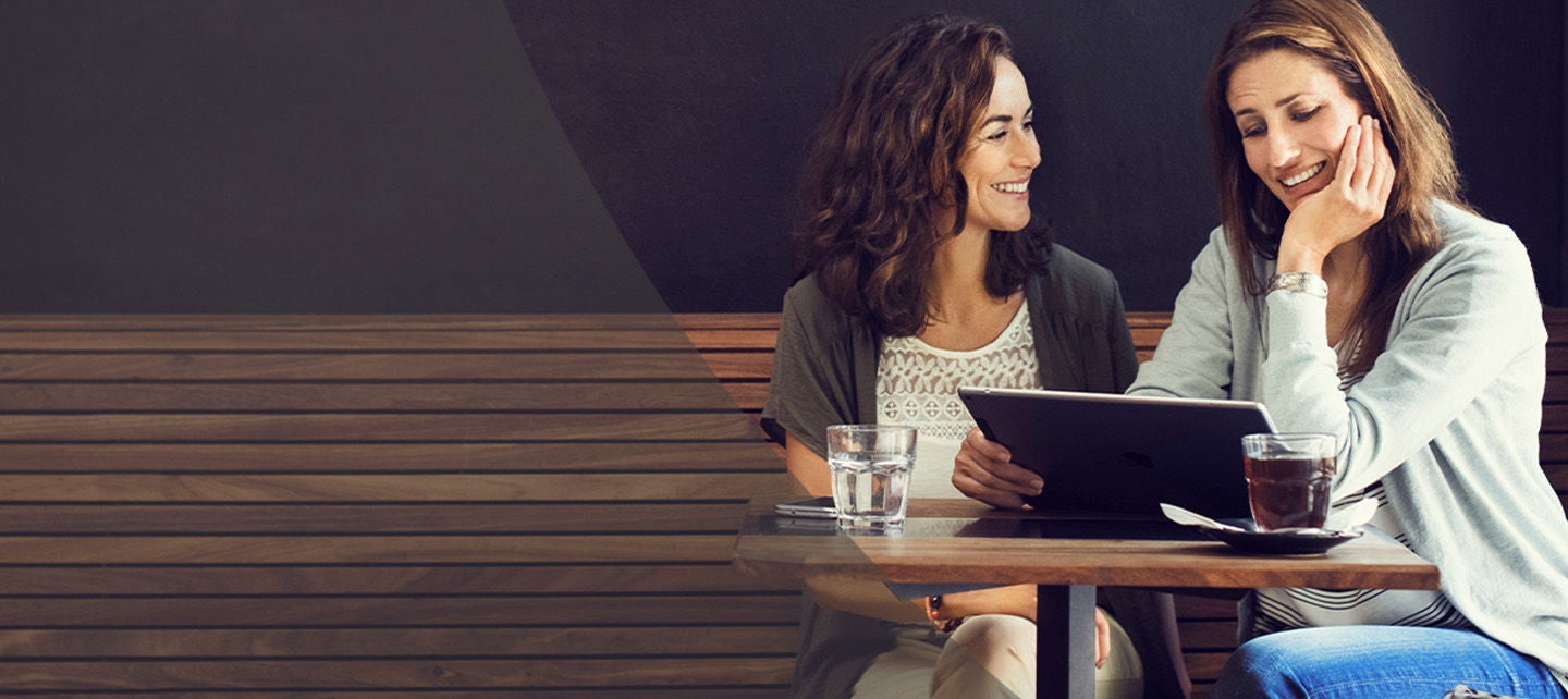 To kvinner ler sammen over kaffe mens den ene viser noe interessant fra nettbrettet.