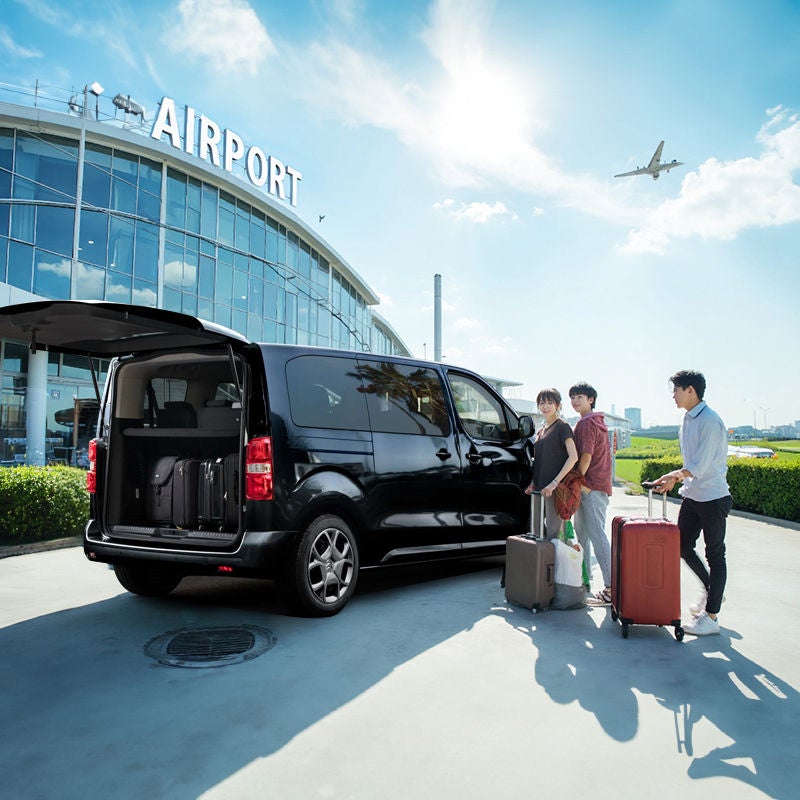 Three travellers with suitcases stand beside a black van with its rear hatch open outside an airport terminal, with a plane in the sky above.