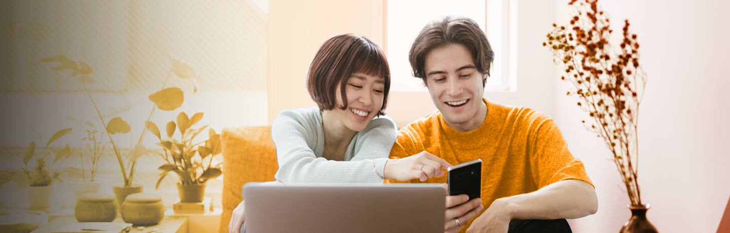 Two people sitting on a sofa looking at a mobile phone with a laptop in front of them