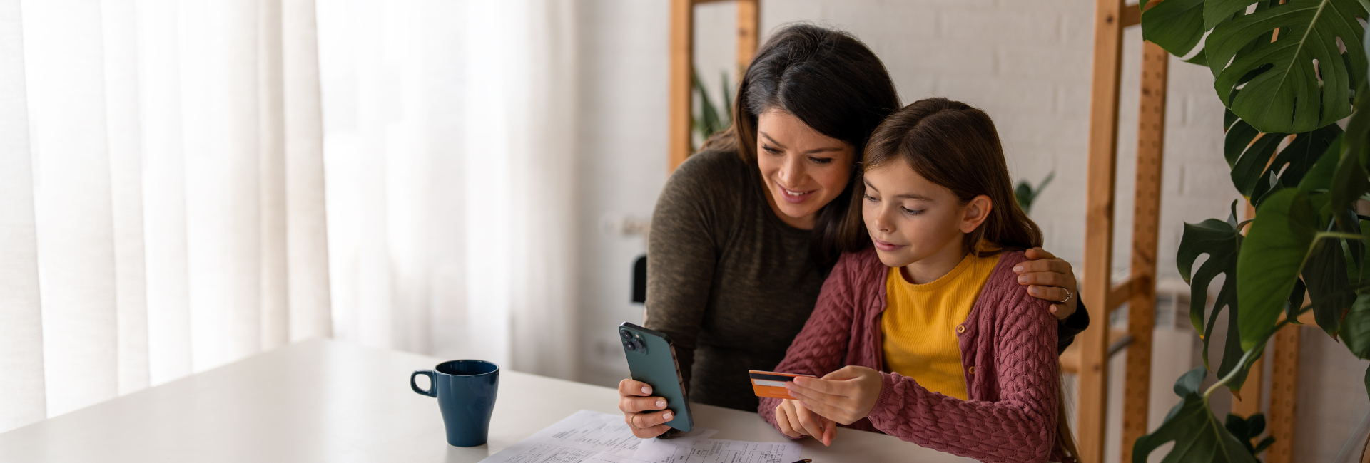 A woman and her daughter look at a payment card and smartphone at a table in a home. 