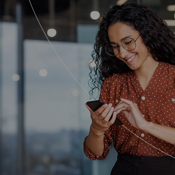 A smiling woman with curly hair and glasses looks at her smartphone while tapping the screen, standing indoors near a window.