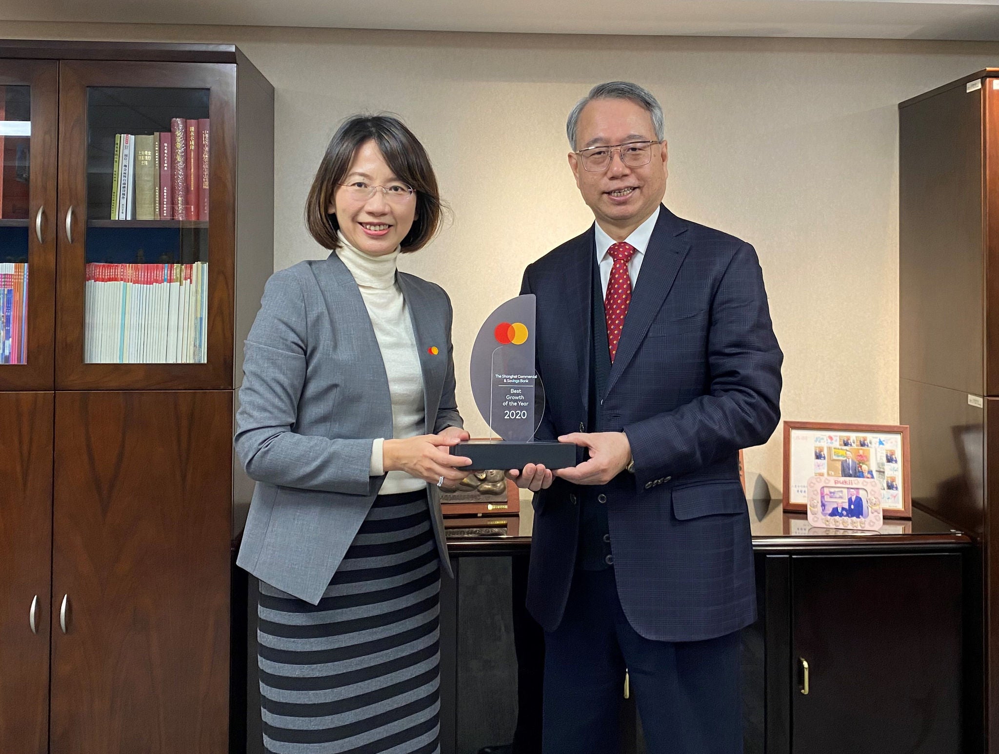 A woman and a man in business attire stand together holding an acrylic award between them in an office with bookshelves behind.