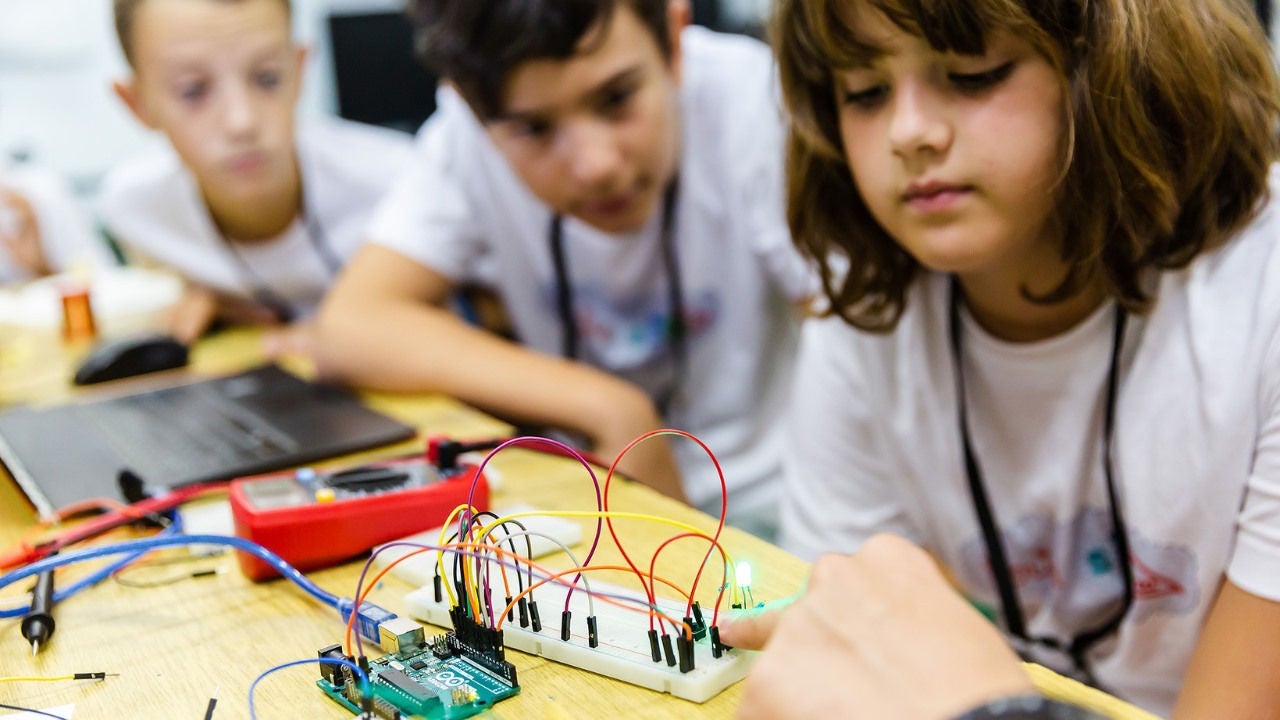 Three children look at a wires attached to a board. 
