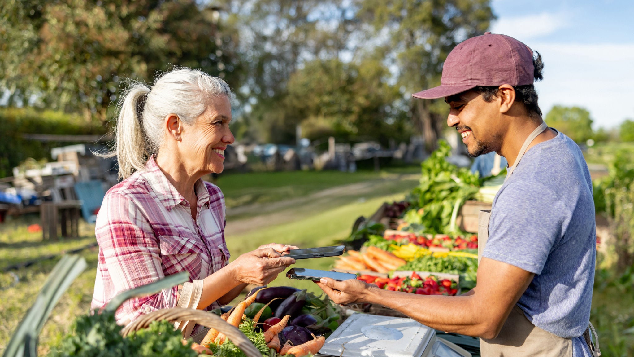 A smiling customer uses her smartphone to make a contactless mobile payment to a vendor holding his phone at an outdoor farmers market stand filled with fresh produce.