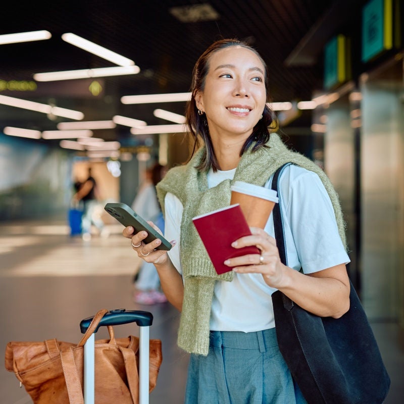 A woman a phone and her passport and a cup of coffee with her suitcase next to her in a transit area. 