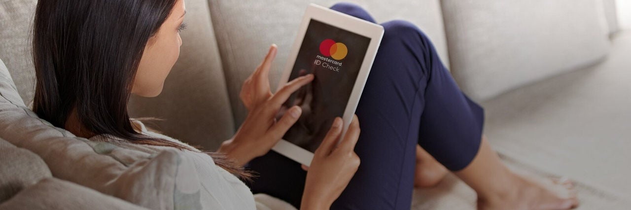 Nő otthon, kanapén ülve táblagépen használja a Mastercard Identity Check szolgáltatást, nyugodt és kényelmes környezetben.