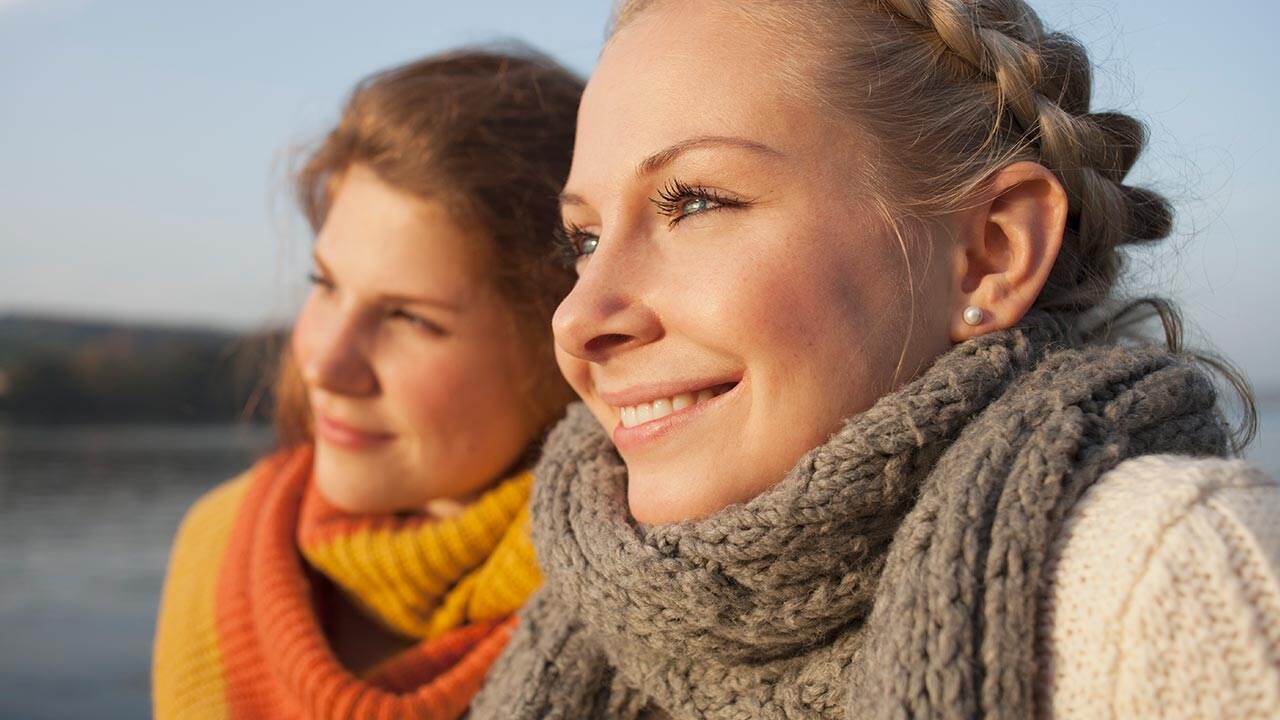 Két fiatal nő meleg sálban, mosolyogva néz a távolba egy tóparton, napfényes szabadtéri környezetben.
