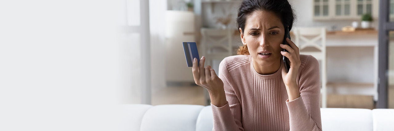 Aggódó nő telefonon beszél, miközben bankkártyát tart a kezében egy nappaliban.