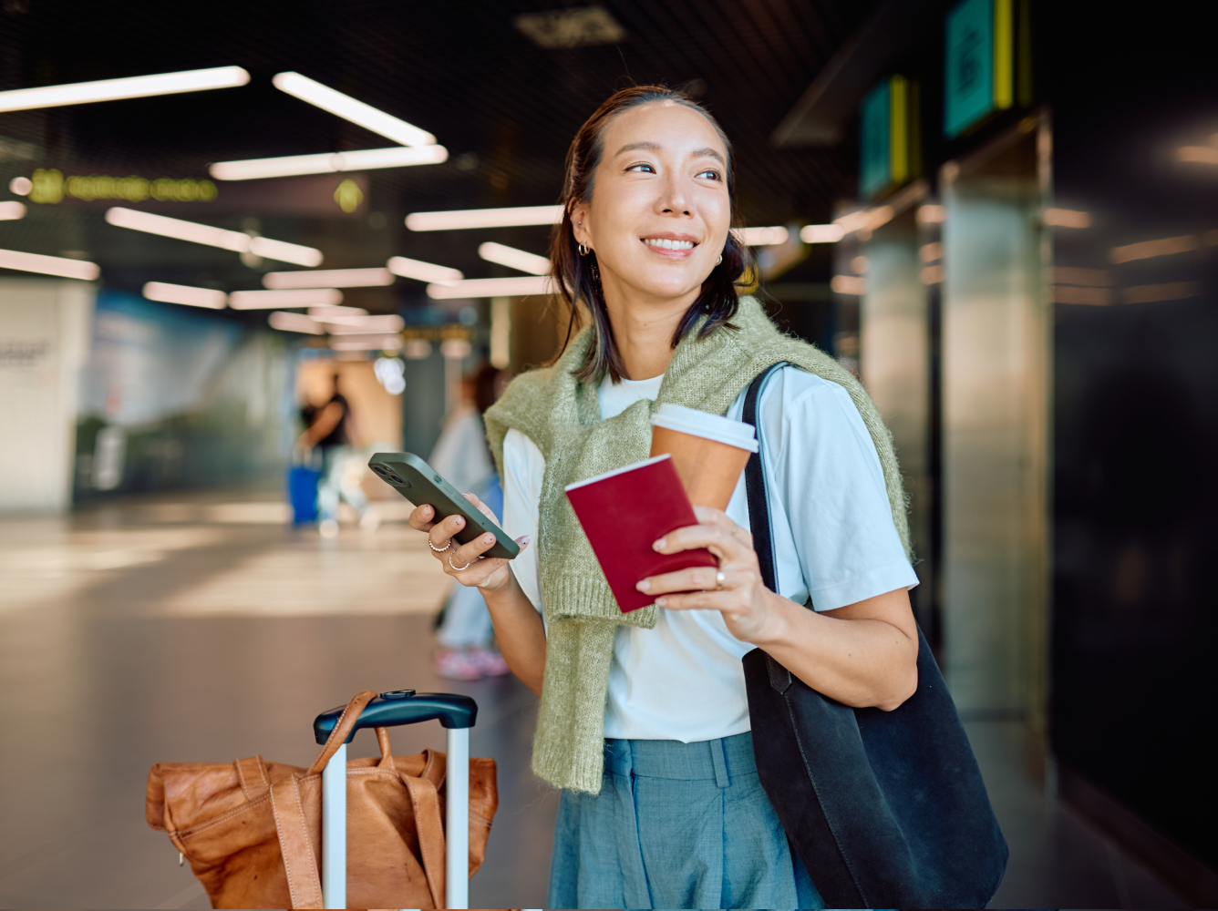 Smiling woman traveler in a bright airport terminal, holding her smartphone, red passport, and coffee cup.