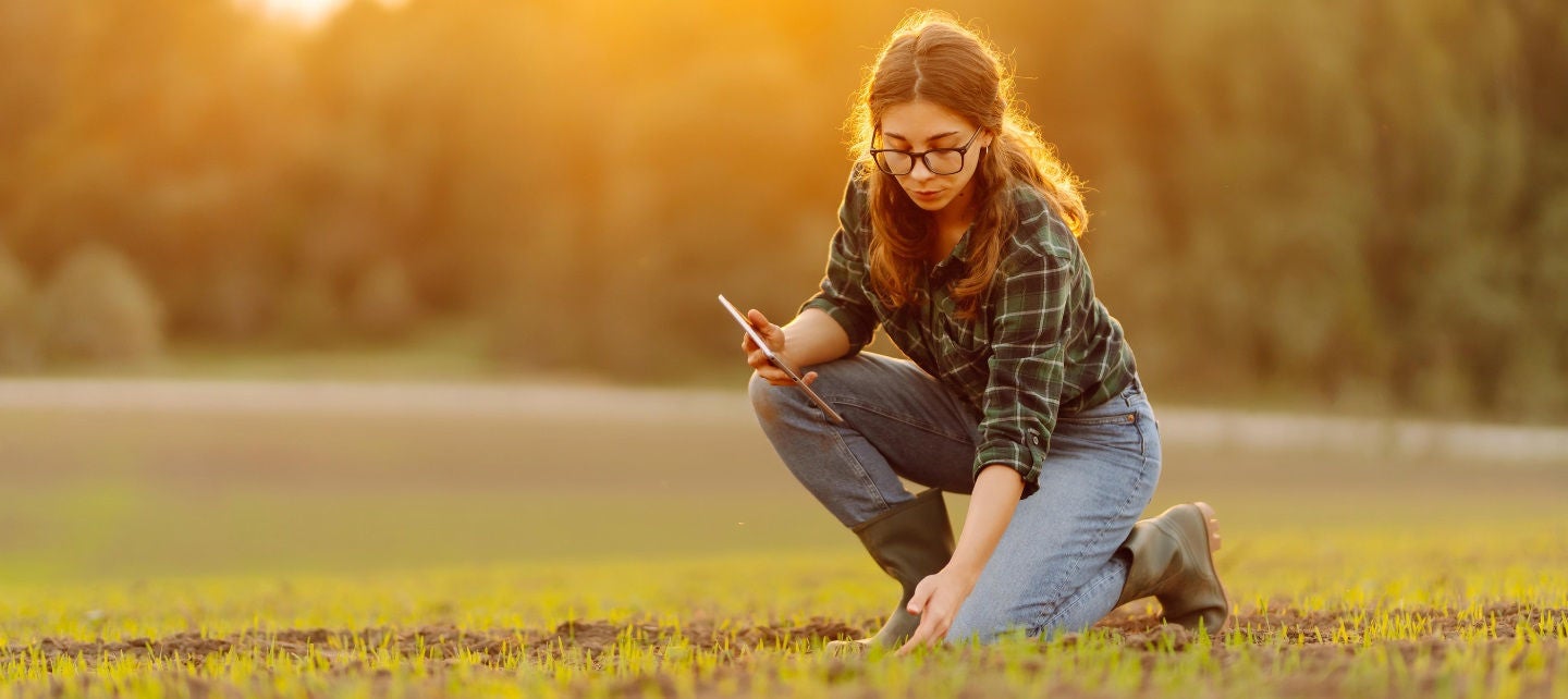 A woman in a plaid shirt and jeans kneels in a field examining sprouts while holding a tablet.