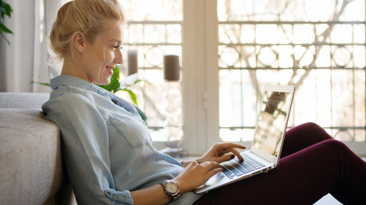 A woman is seated on a couch in her home smiling at her laptop screen.