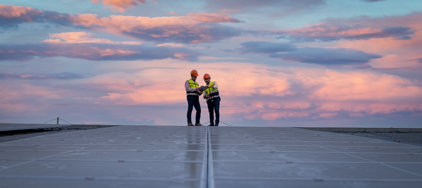 Dois homens com coletes de trabalho estão em pé no topo de um campo de painéis solares ao nascer do sol. 