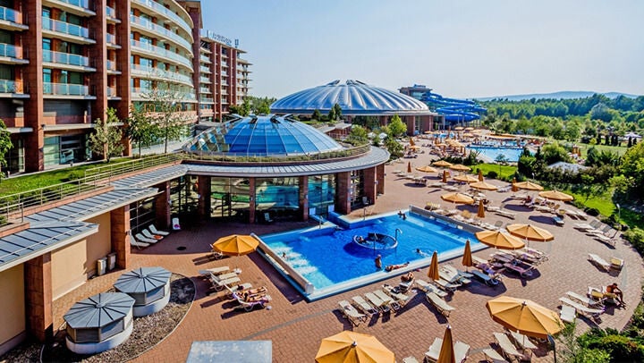 Szabadtéri medence wellnesskomplexumban, napozóágyakkal, napernyőkkel, körülötte modern szállodaépületek és zöld terület