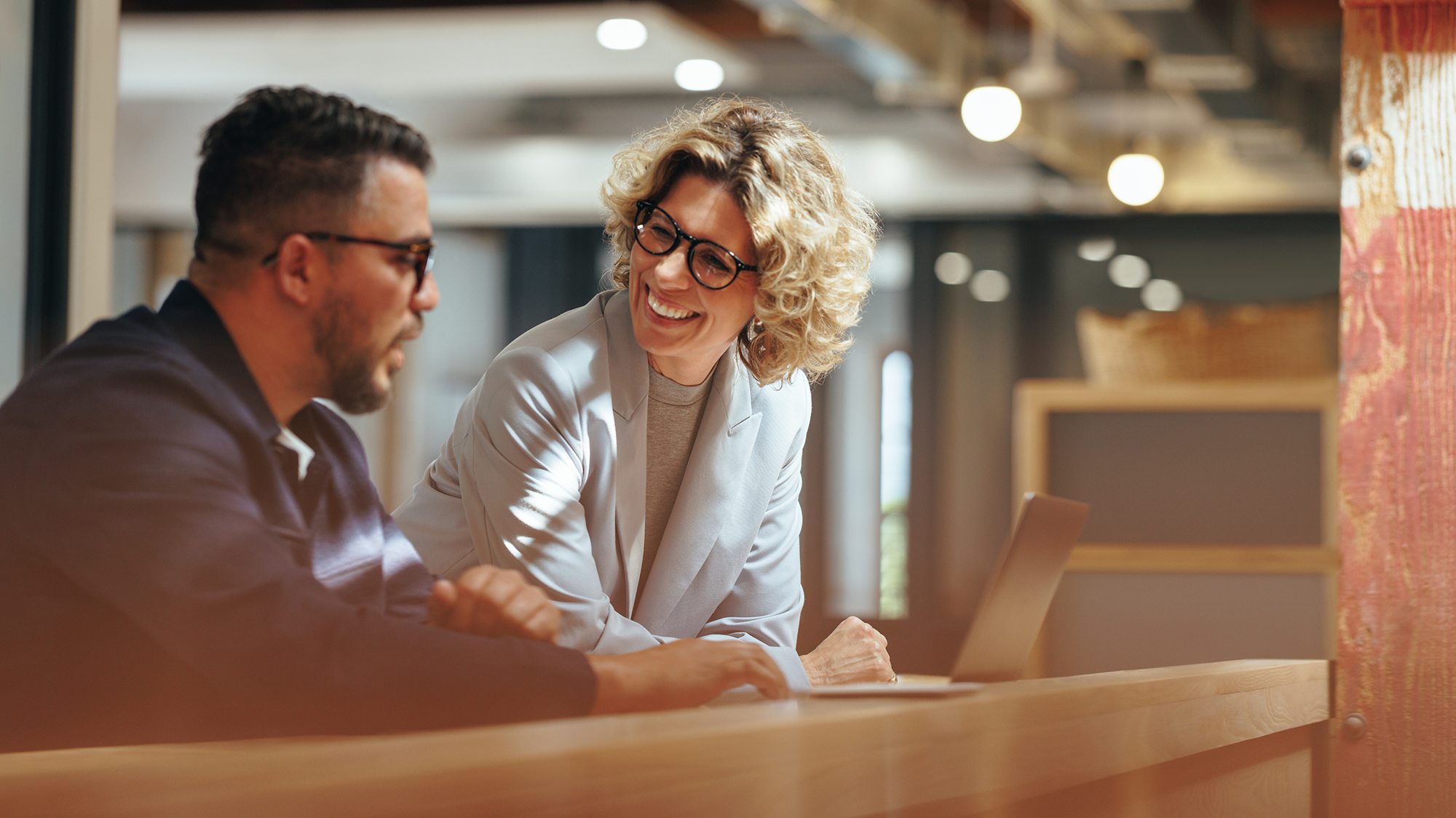 Two colleagues, a man and a smiling woman, working together at a laptop in a modern office environment.
