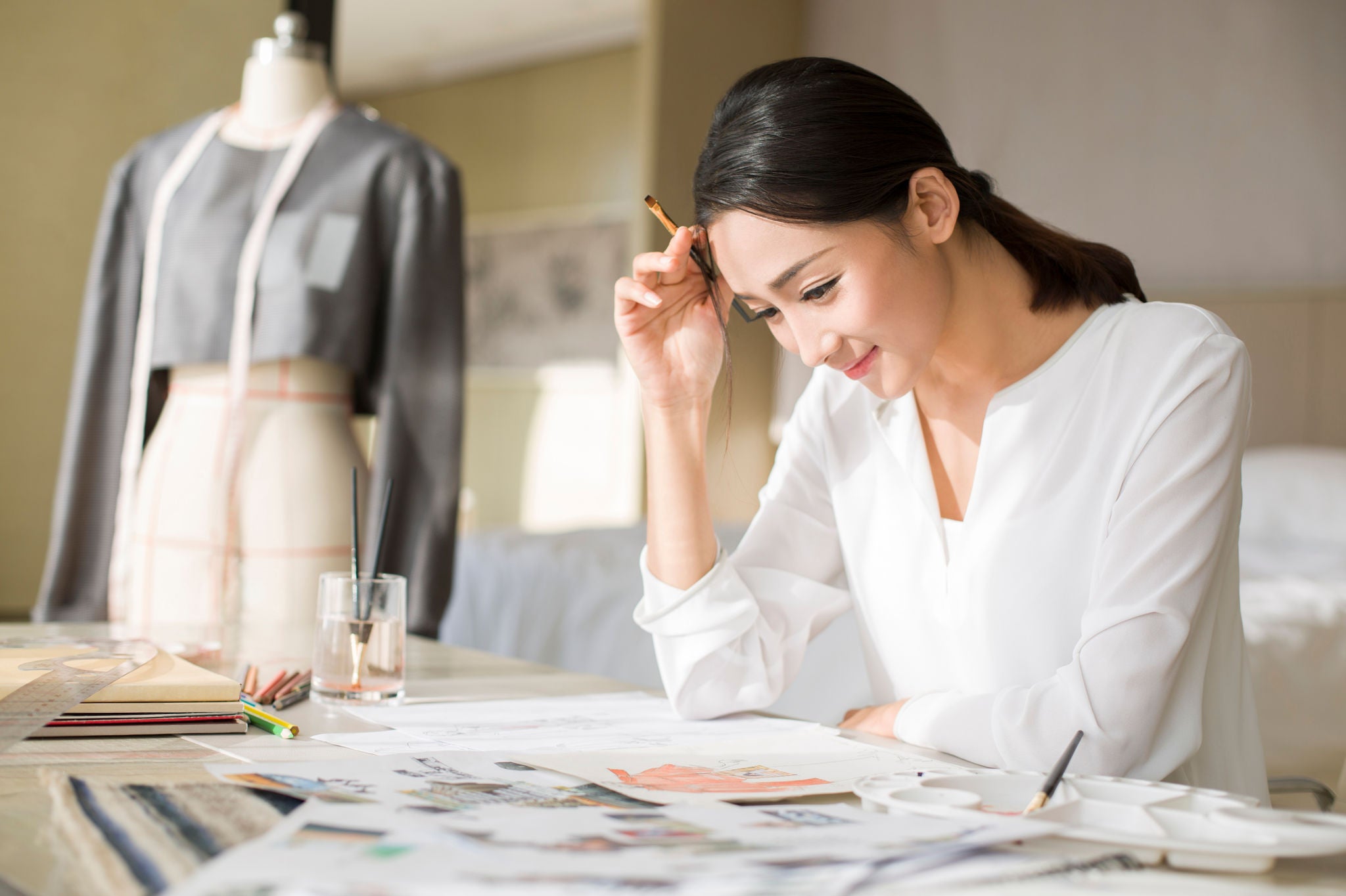 A woman in a white blouse sits at a desk sketching designs, with a dress form and drawing materials around her.