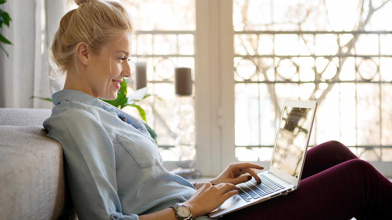 Egy mosolygó nő otthoni környezetben, egy kanapén ülve laptopon dolgozik természetes fényben.