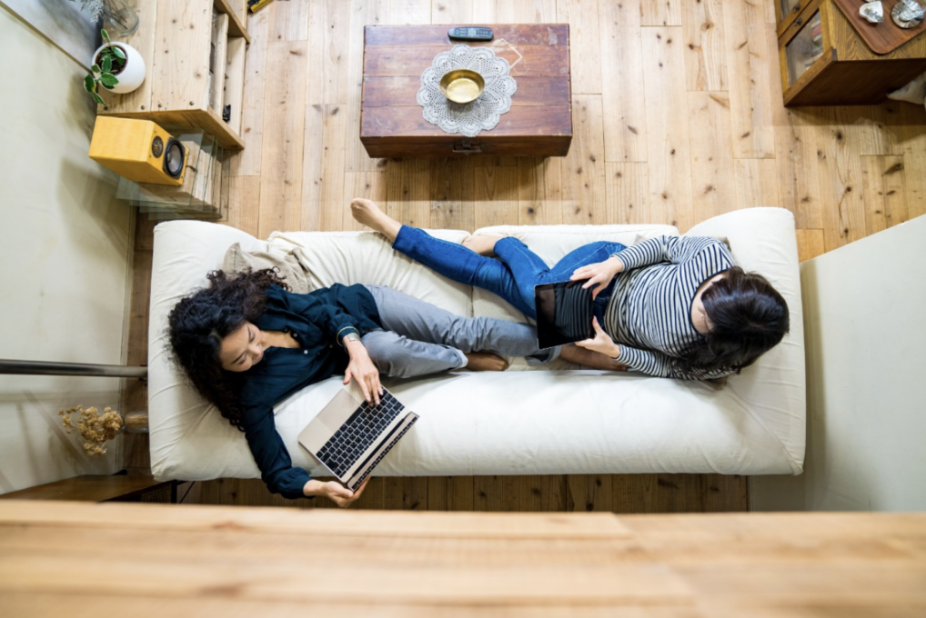Zwei Frauen sitzen auf einem Sofa und schauen auf einen PC und ein Tablet.