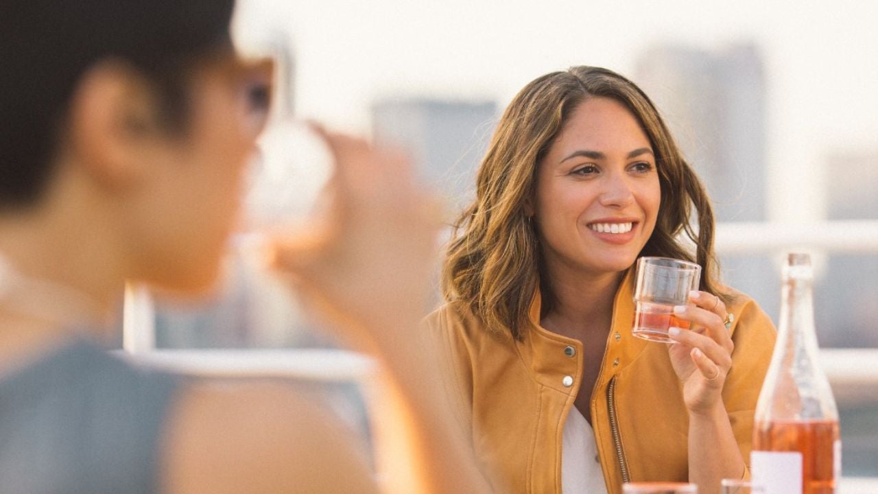 Egy nő napfényes teraszon rosé bort kortyol, miközben beszélget valakivel; az előtérben egy másik személy elmosódva látható.”