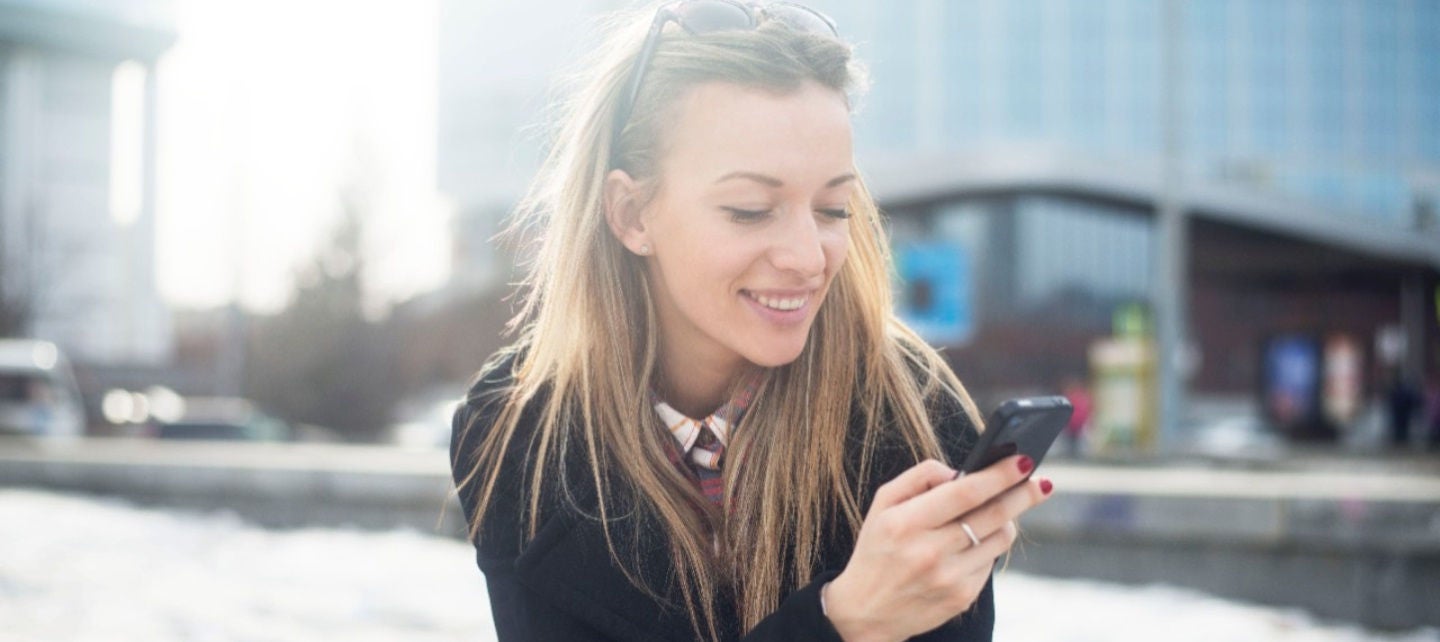 A younger woman sitting outside next to an office building looks at her smartphone.
