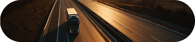 A large white truck drives on an empty highway during sunset. The sky is partly cloudy with warm tones reflecting on the road..