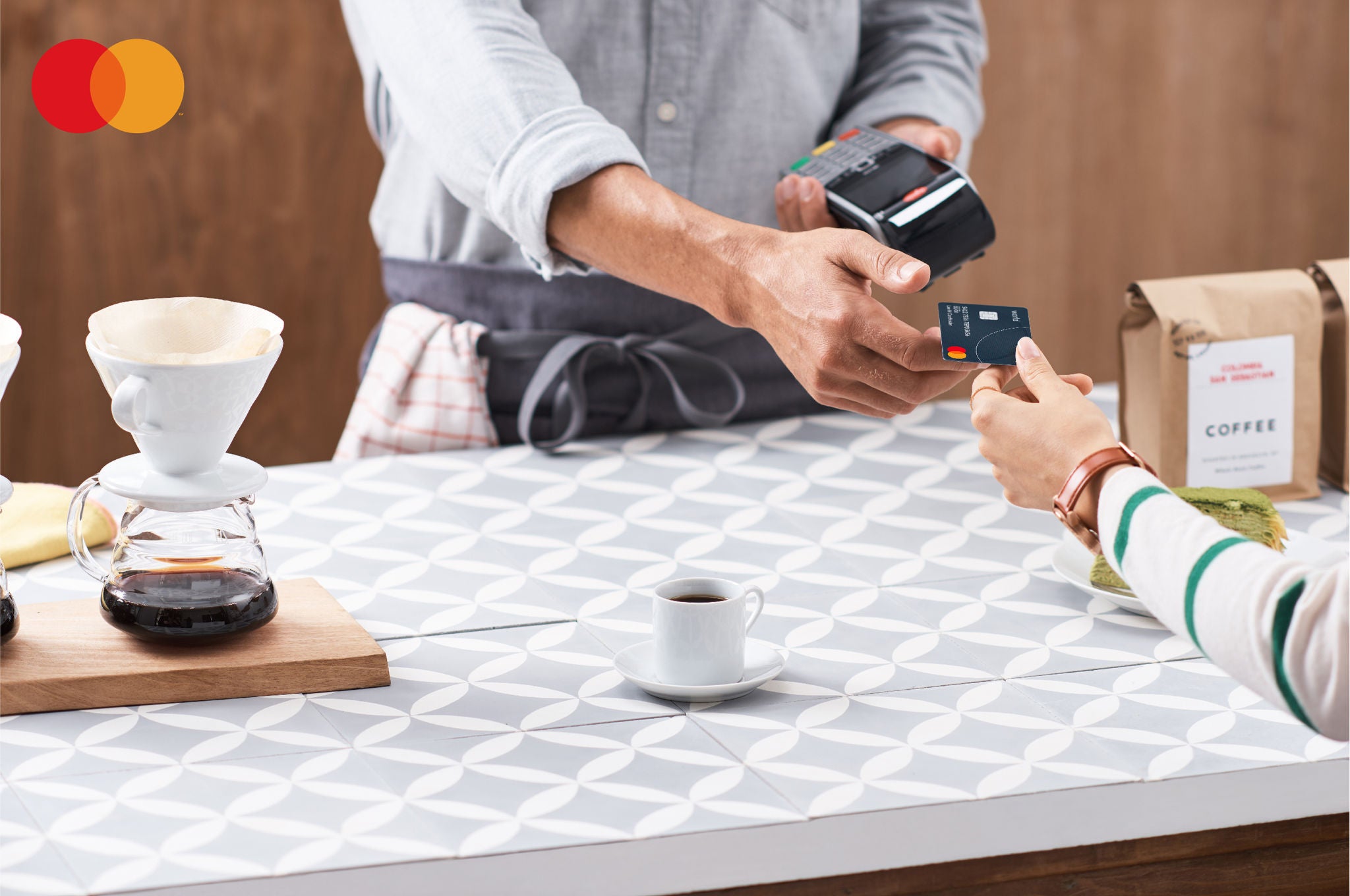 A close-up of a person in an apron holding a card terminal while a customer hands over a payment card across a café counter, with coffee equipment nearby.