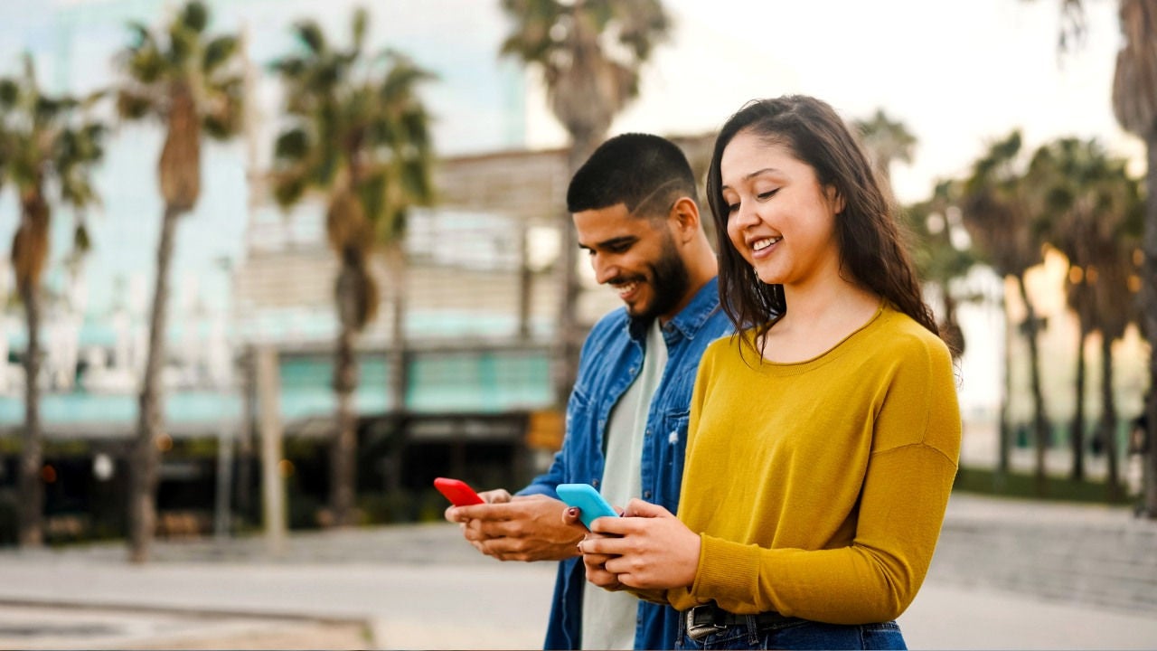 A couple look down at their phone while walking ina tropical outdoor plaza. 
