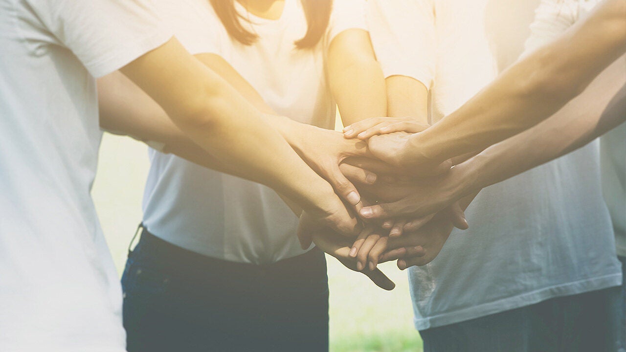 Több ember összetett kezei fehér pólóban, csapatmunkát, összetartást és közös célért való együttműködést szimbolizálva.