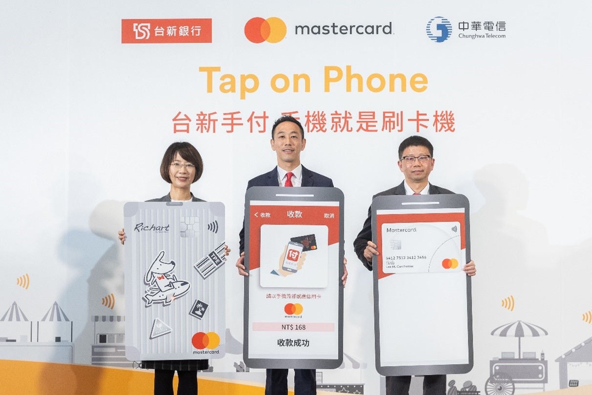 Three people in business attire stand on stage holding oversized cutouts of a payment card and phone screens, in front of a backdrop with brand logos.