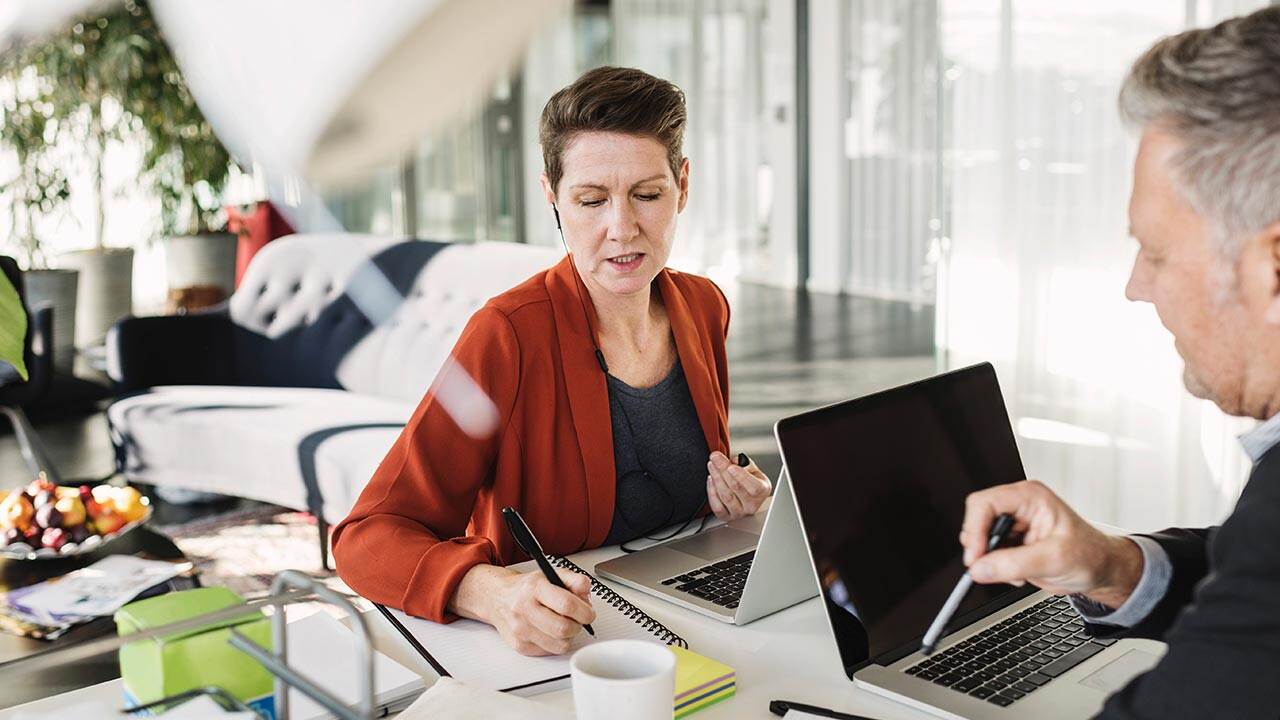 Két kolléga laptopokon dolgozik egy modern irodai környezetben, jegyzetelés és megbeszélés közben egy asztalnál