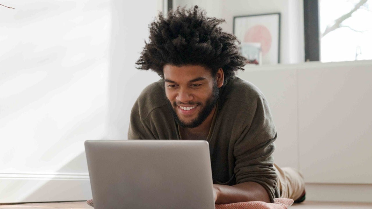  A man smiles stretched out on the floor of an apartment smiles at his laptop