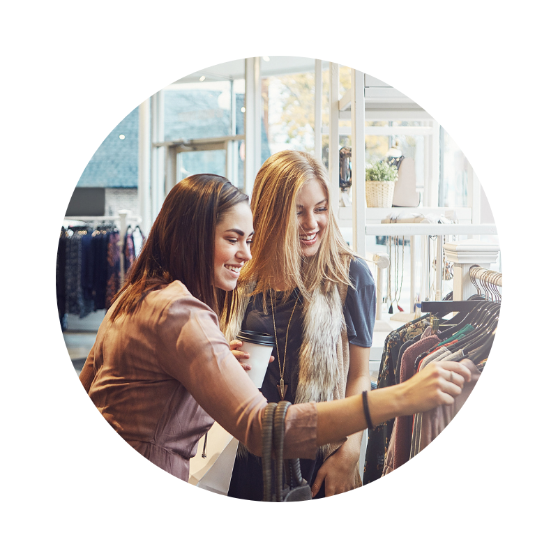 Két fiatal nő ruhaboltban vásárol, mosolyogva válogatnak a ruhák között egy állvány előtt, baráti és élményszerű vásárlási helyzetben.